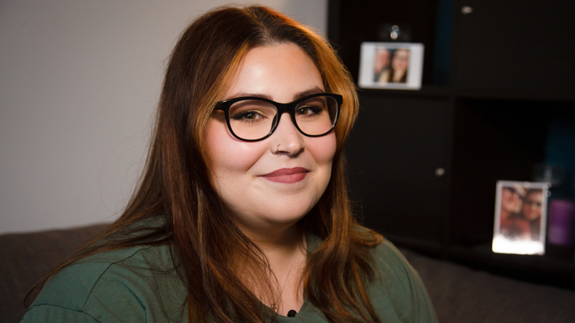 Holly is looking at the camera. She is wearing black framed glasses and is wearing a pink blush lipstick. Her brown hair is untied and frames her face, she has two blonde streaks at the front. She is wearing a khaki green t-shirt and in the background there are framed photographs. They are slightly blurred as the camera focus is on the woman.