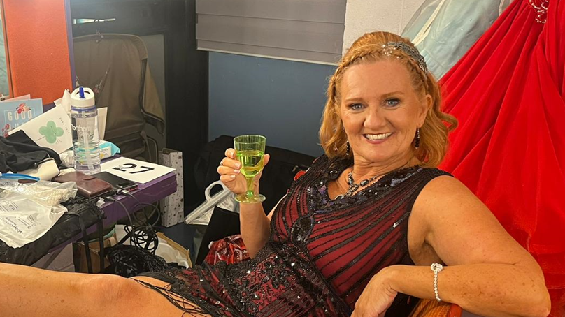 A woman in dance costume relaxes backstage. She is reclining with her feet up and is drinking from a green-coloured glass. She has blonde hair and is smiling.