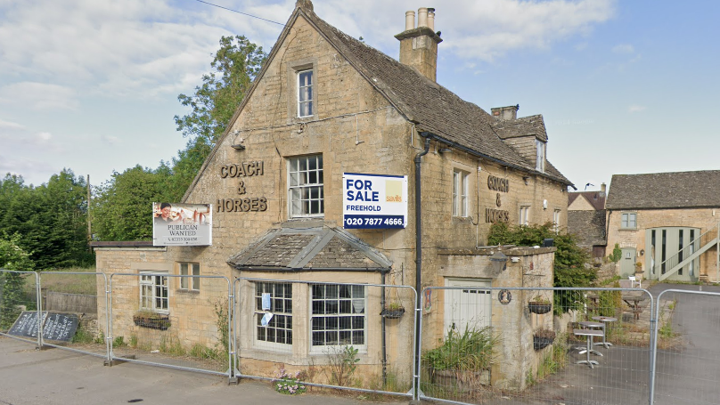 A closed pub in the Cotswolds, made of Cotswold stone. It has a large bay window on the ground floor and is surrounded by metal construction fencing. It has signs that read "for sale" and a metal sign that reads "Coach & Horses".
