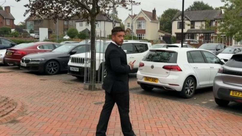 A man in a black suit looks over his shoulder at the camera as he walks toward a row of parked cars in a car park with a row of houses visible in the background