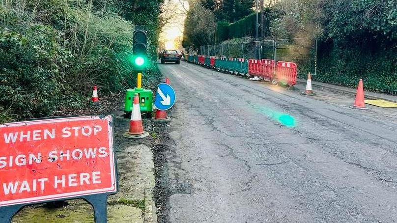 Temporary traffic lights near a partial road closure in Mickleover in Derby. Temporary fencing is in place against a wall on the right side of the road.
The light is on green and a car can be seeing travelling away from the camera in the distance.
A sign in the foreground of the image reads 'when stop sign shows wait here'.