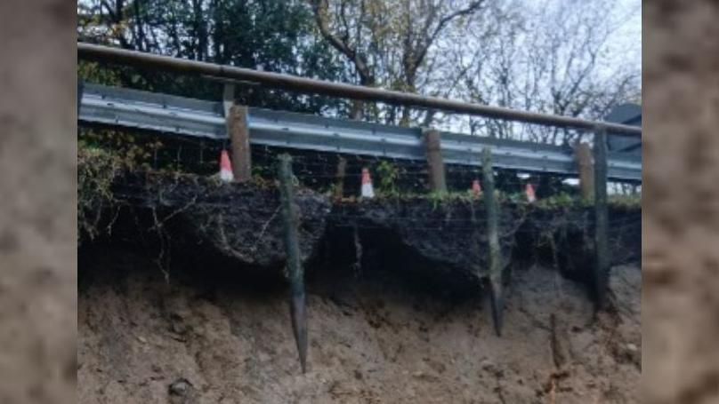 Close up shot of the side of the road with a grey fence in the foreground and trees in the background. There are orange cones by the fence too. Underneath the fence is exposed soil which shows signs of a large landslip. 