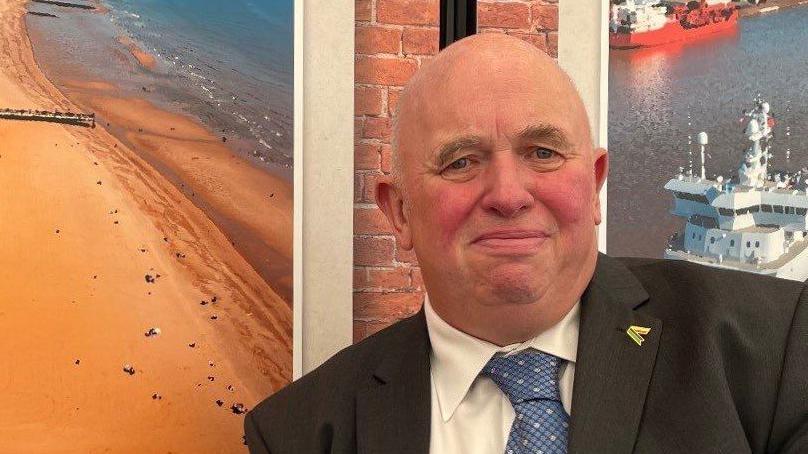 Councillor Colin Davie, wearing a dark suit and blue tie, sitting in front of a poster advertising Skegness beach