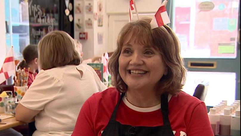 A woman in a red top is smiling widely ad the camera, she has a head band with two England flags on sticks. In the background a women in a white top has her back to the camera and a girl with a ponytail is looking away from the camera