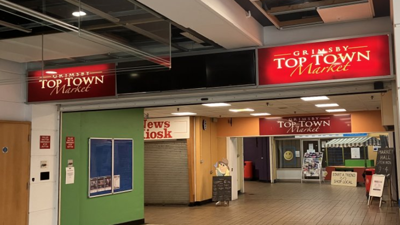 The entrance to Top Town Market. There are three signs overhead bearing the market's name.  A News Kiosk sign can also be seen. The kiosk has its shutters down. 