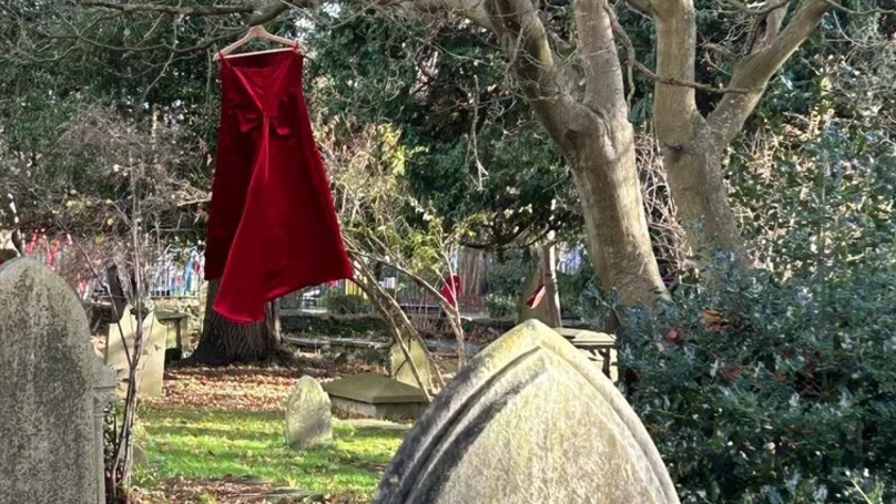 A red velvet dress hanging on a hanger from a tree in a churchyard with the tip of a large gravestone in the foreground