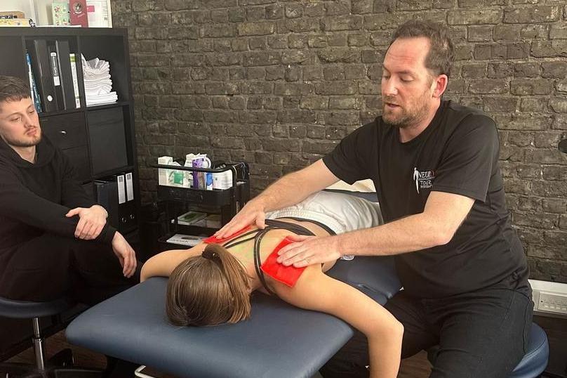 A man massages a woman lying on a massage table in a brick studio while another man looks on.