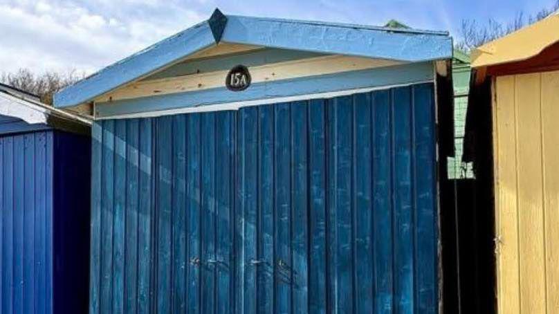 The beach hut before the renovation. It has navy wooden boards blocking its entrance. The top has panels that are light blue-and-white striped. The roof is light blue.