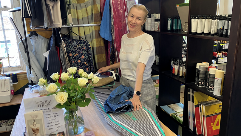 A woman stands smiling behind an ironing board. There are racks of clothes behind her and a vase of white roses to one side. She is holding an iron in one hand and a pair of jeans with the other.