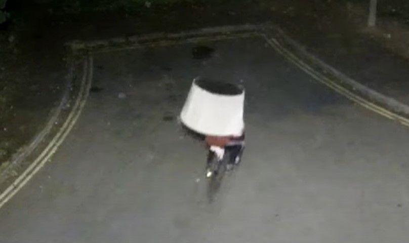 A man riding a bicycle whilst balancing a large white object on his head. It is unclear if the object is a lampshade or an upturned chair.