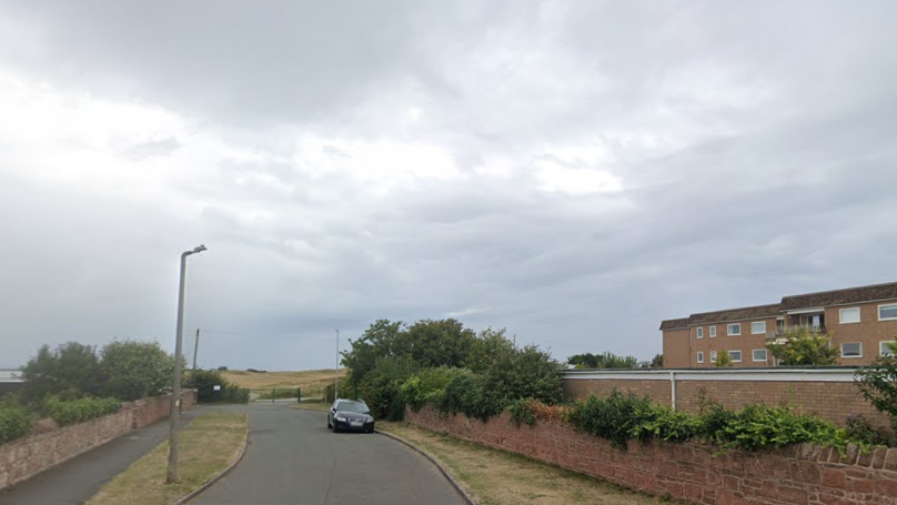 A cul-de-sac called Pinfold Lane. It is a short road leading to a gate, through which is the course of the Royal Liverpool Golf Club. There are low walls on either side of the grass verges, and a black Audi is parked on the right.