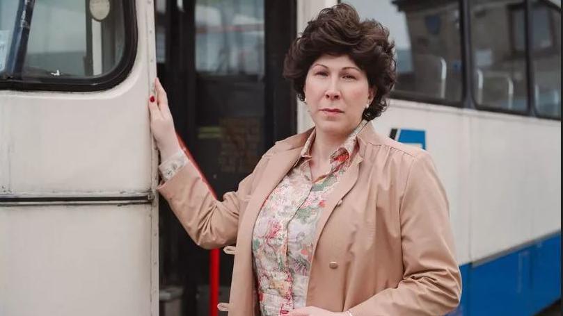 A woman wearing a beige coat and floral blouse stands at the door of an white bus. 