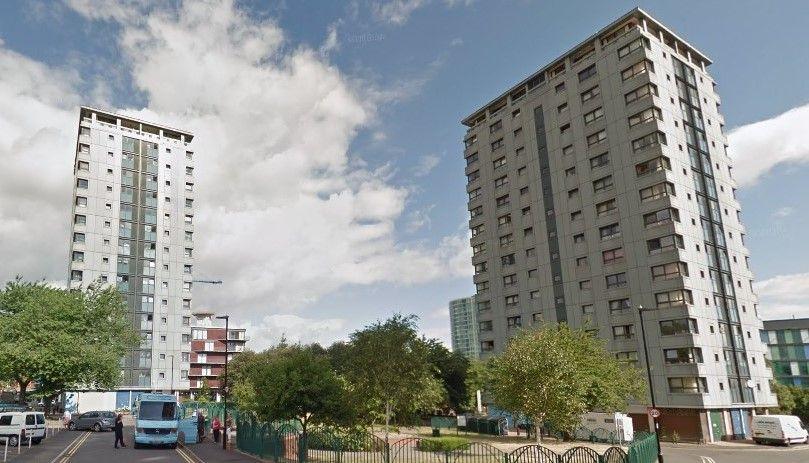 Grey blocks of flats under a blue sky with fluffy clouds. At the foot of the flats are several trees and a small fenced-off formal garden.