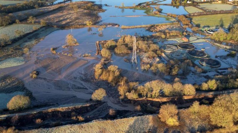  A drone shot of a canal which has burst in one section with floodwater and sediment running into surrounding fields. 