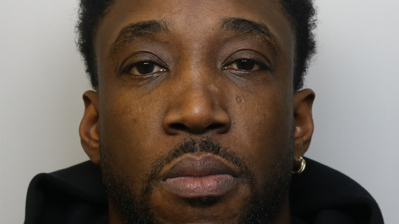 A man looking at the camera in a police mugshot, with a goatee and wearing a hopped earring and a black hoody.