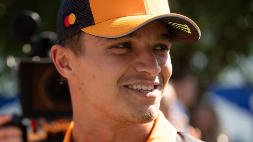 Lando Norris smiles while wearing a McLaren baseball cap with a camera filming him from behind