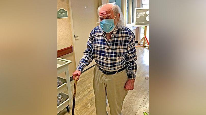 An elderly man wearing a plaid shirt and a surgical mask stands with the assistance of a cane