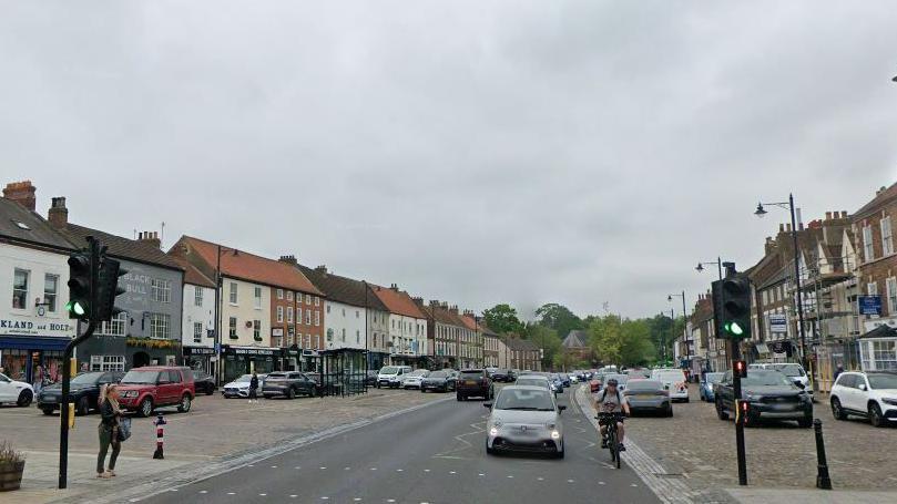 Google Streetview of Yarm town centre. A number of vehicles are travelling along a main road near traffic lights. Other vehicles are parked to either side of the road.