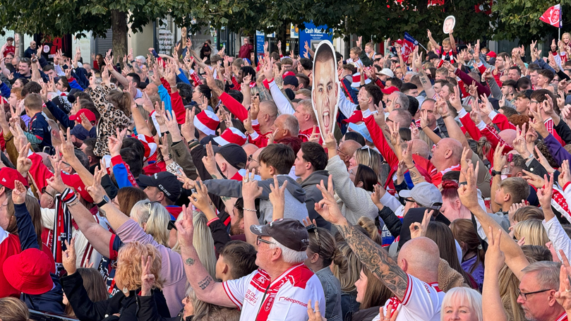 A wide shot of the crowd with them all holding up one arm with three fingers showing.  They are mostly dressed in red and white clothing.