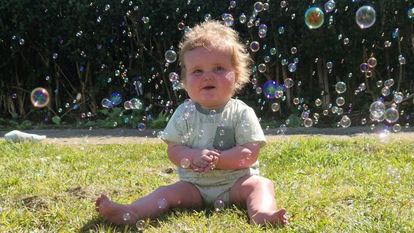 A baby with curly ginger hair in a pale green top and a nappy is sitting on grass, with bubbles floating all around him.