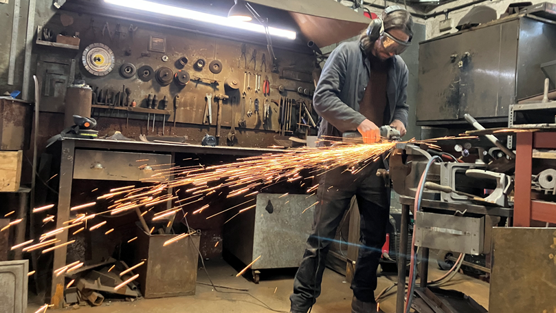 Olly Osborne, wearing goggles, is operating a metal grinder. Sparks fly across the room spectacularly. Behind him is a rack of metal-working tools and other equipment used to restore ovens.