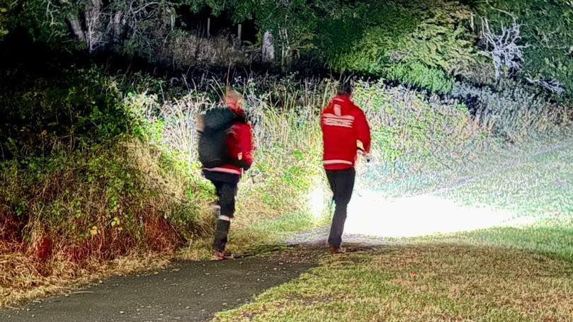 Two mountain rescue volunteers - wearing bright red emergency clothing - walk into a wooded area. It is late evening and the volunteers are using torches to light up their route. 