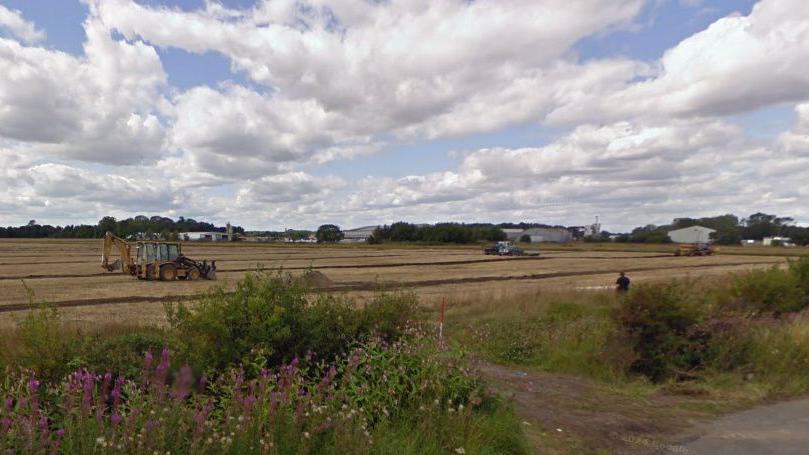 A field which is the site of the proposed solar farm. Three farm vehicles are in the field. 