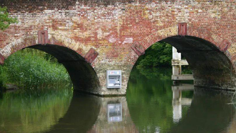 Two arches of the red brick bride reflect in the river with the grey atm attached to a central buttress