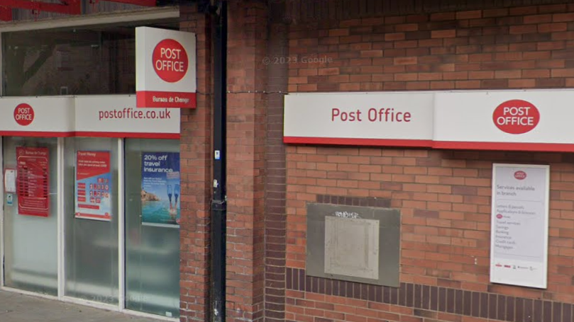 Photo shows the outside of Rotherham town centre post office. There is a shop window and brick wall, with red and white Post Office signs