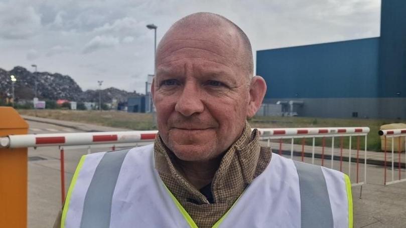 A bald man wearing a silver high-vis vest stood in front of barriers to a car park