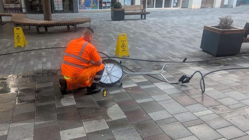A workman in orange hi vis connects a piece of cleaning equipment in a pedestrianised area, with a planter containing a shrivelled shrub to the top right of the picture.