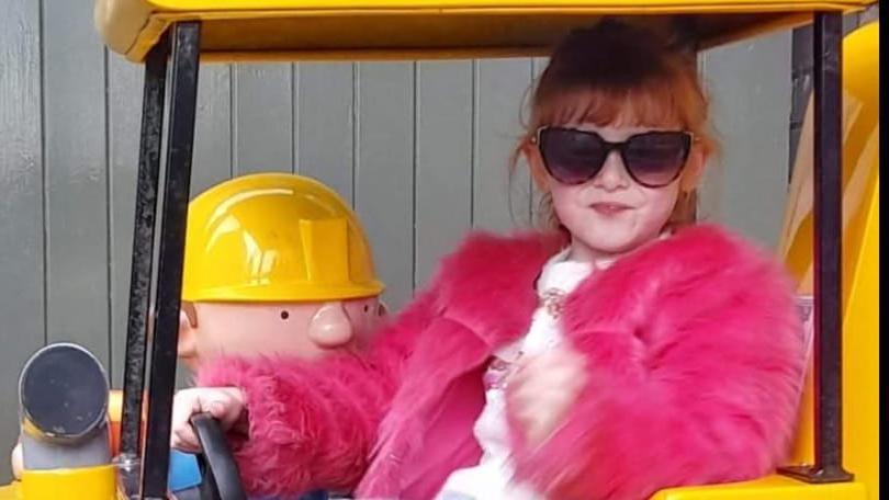 Lila is sitting in a yellow tractor and has one hand on the steering wheel. She is wearing a pink fluffy jacket and has sunglasses on.