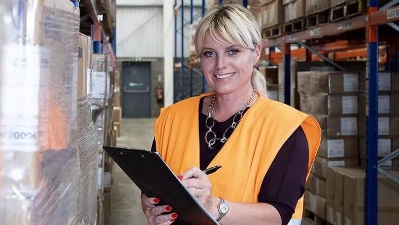 A smiling Sarah Stoute looking directly at the camera holding a clipboard and pen, and wearing a orange high-visibility jacket.