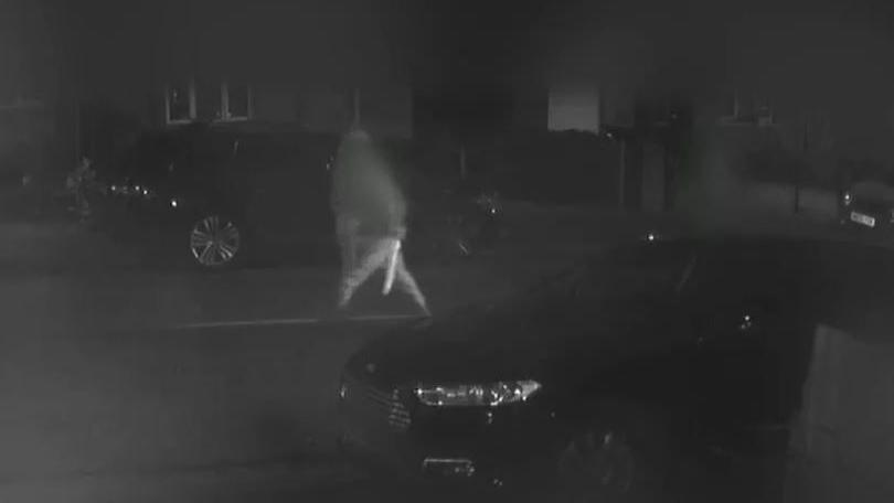 A black and white screenshot of CCTV footage from a house opposite the street. It shows one of the defendants walking across the road, carrying a large machete which is glinting in the streetlights.