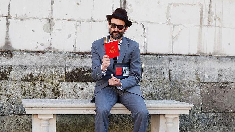 Mark Oliver Everett sits on a stone bench wearing a grey suit, hat and sunglasses. He is reading an open red book and has his Freedom document tucked under his arm