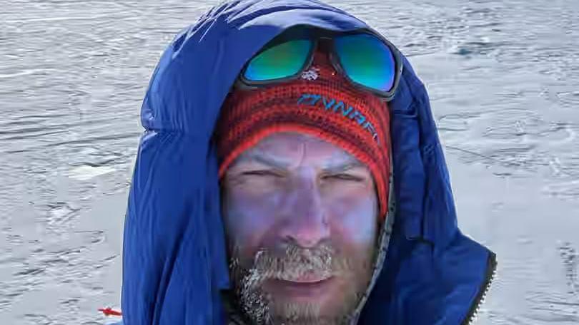 Jonny Huntington wears a blue coat with its hood up and a red woolly hat while stood in the Antarctic. He has a blonde beard which has signs of frost in it. He has a pair of sunglasses perched on his forehead.