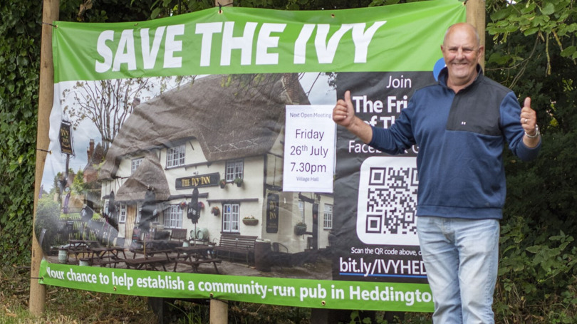 Paul is standing in front of a hedge beside a large banner saying 'Save the Ivy', which has a picture of the pub on it. He is holding both thumbs up and smiling to camera