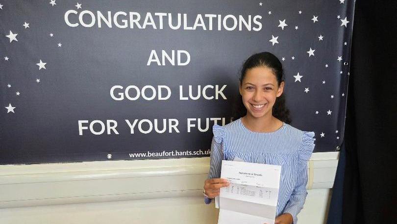 Élise is standing in front of a congratulations sign, holding her exam results on a piece of paper. She has black hair, tied back in a ponytail and wears a blue and white striped, frilly long sleeved blouse. She is grinning widely for the camera. Behind her is a navy blue sign with white stars printed on it and the writing "Congratulations and good luck for your future"