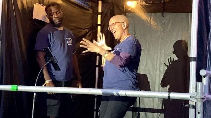 A woman who is doing sign language standing in a gazebo next to the main stage at a festival. She is wearing black jeans and a navy top. A man is also standing to her side.