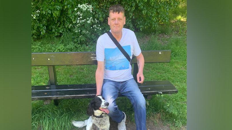 Steven Mead wearing a white T-shirt with a light blue square at the front of it and blue jeans. He is sitting on a park bench and smiling at the camera.
He has a dog sitting next to him.