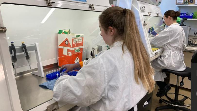 Two women wearing white boiler suits sitting in front of a row of test tubes in a laboratory.


