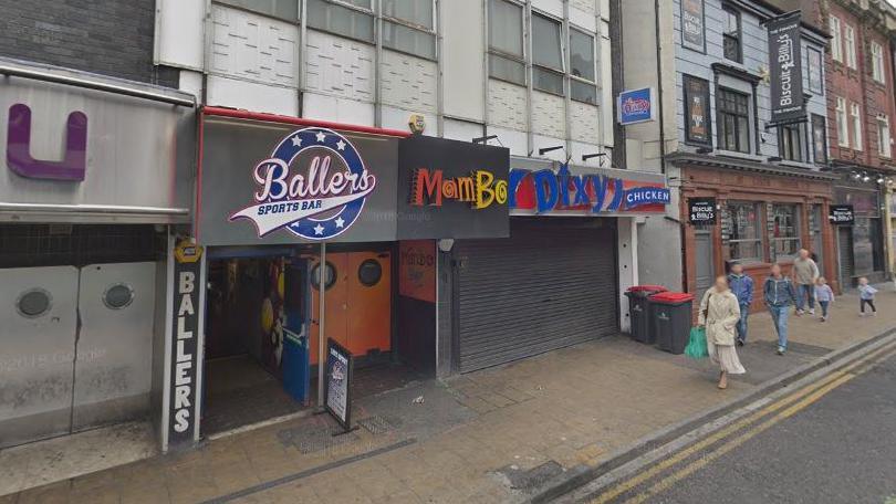 The club is located between Ballers Sports Bar and Dixy Chicken. The letters of the club's sign are in red, orange and yellow. Four adults and two children can be seen walking on the pavement outside the club.