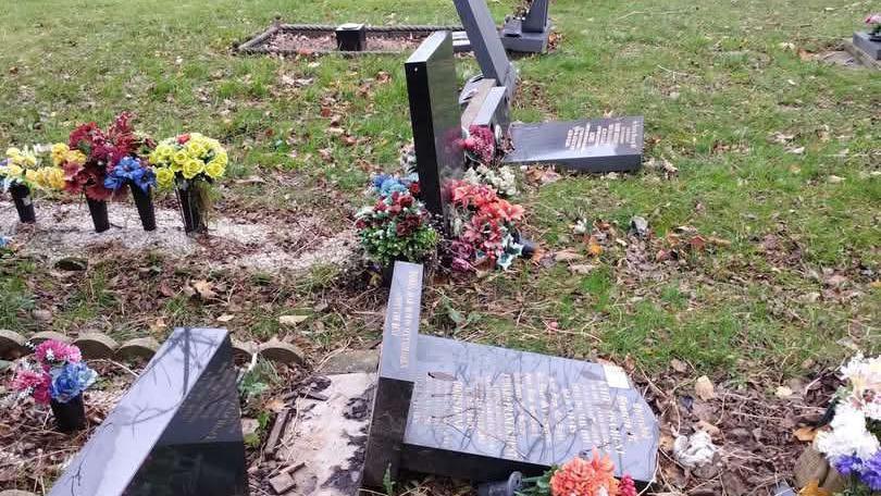 A close-up view of a row of graves in a churchyard, showing multiple headstones that have been knocked over or are leaning at sharp angles. The graves are adorned with artificial flowers in bright colors such as red, yellow, and pink. The ground is covered with leaves, and the stone church building with arched windows and a doorway is visible in the background.