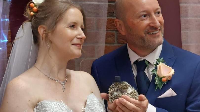 A photo of Michelle and David Doughty at their wedding. They are facing away from the camera and smiling. Mr Doughty is holding Pico the hedgehog in his hands while Pico has a small black top hot on his head.