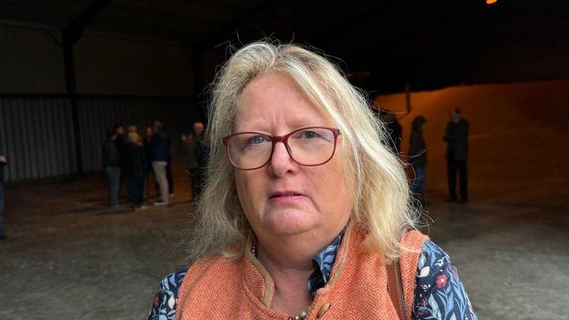 A woman looking straight at the camera. She is wearing red glasses, a blue top with floral print, and an orange gilet. Several people are standing in a building in the background.