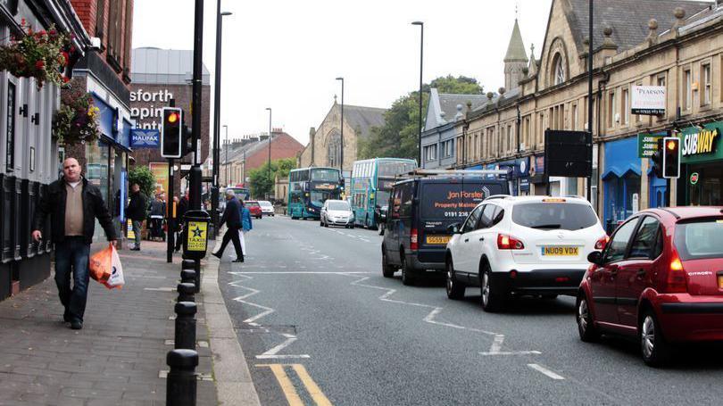 A high street with a queue of cars waiting at traffic lights in the middle lane. The left lane is empty.