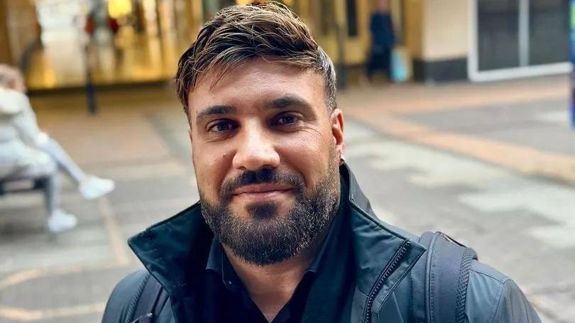 Alastair smiling at the camera. He is wearing a dark puffer jacket and has dark facial hair and short brown hair.