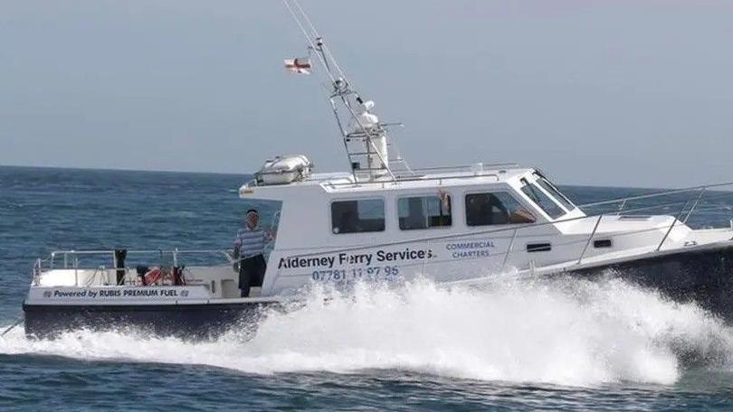 A man in a blue striped shirt driving on a small white ferry on water. There is water splashing up the side of the ferry.  On the side of the ferry it has various words and numbers including Alderney Ferry Services