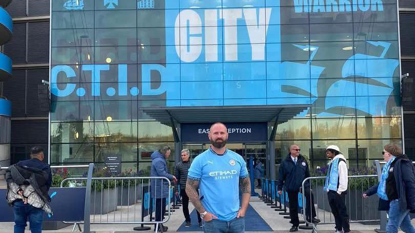 Manchester City fan Aaron Cowley stands outside the Etihad Stadium wearing a sky blue Manchester City shirt. Several other people are walking around near him. He has a number of tattoos on his arms and is wearing blue denim jeans.