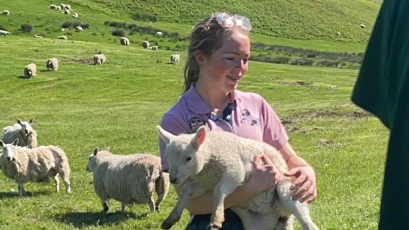 Kaitlyn Singer, who is 16 and wearing a pink top with glasses on her head is holding a very fat lamb. Behind her the hills are full of grass 